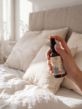 Verpackungsdesign für die Selfcare-Marke Grinsekatzen – Hand hält ein CBD-Spray der Sorte Pur vor einem gemütlichen Bett mit hellen Kissen.